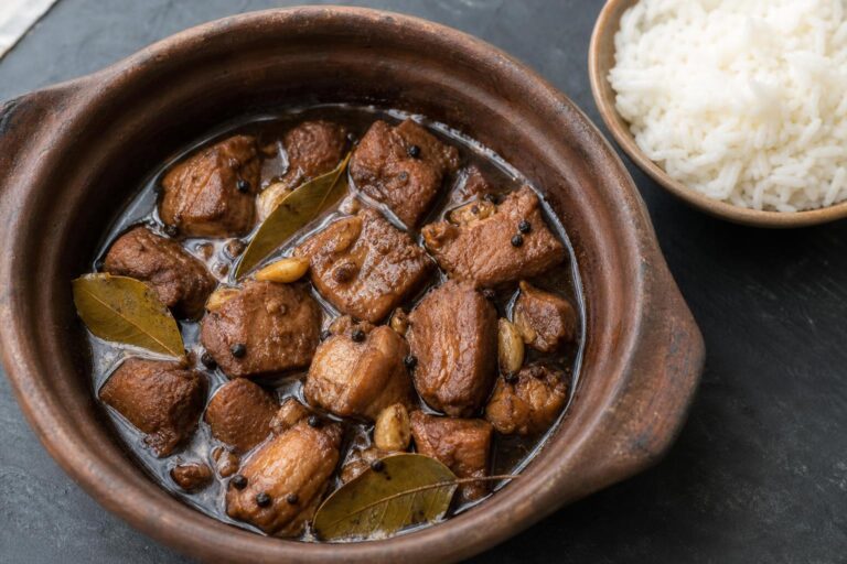 Pork adobo with bay leaves and garlic in a clay pot next to a bowl of rice