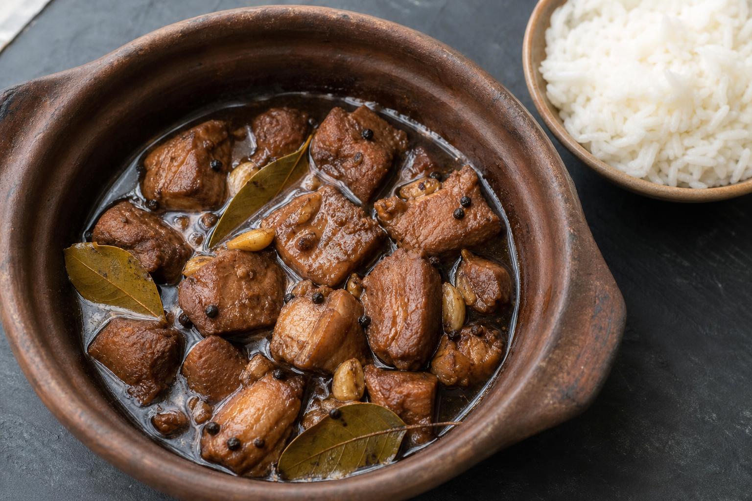 Pork adobo with bay leaves and garlic in a clay pot next to a bowl of rice
