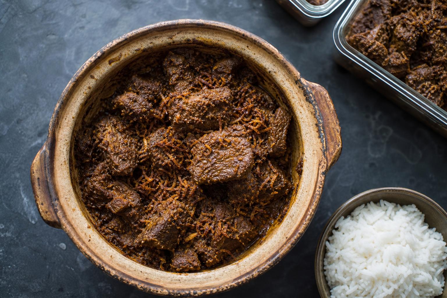 Beef rendang in a clay pot with rice and meal prep containers alongside