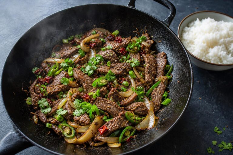 Cumin beef stir fry with seared flank steak, onions, green chili, and toasted cumin seeds in a cast iron wok with a bowl of rice