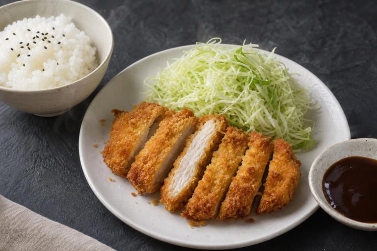 Sliced air fryer chicken katsu on a white plate with shredded cabbage, tonkatsu sauce, and a bowl of rice