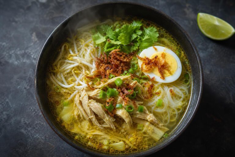 Overhead bowl of soto ayam, golden turmeric chicken soup with shredded chicken, noodles, bean sprouts, egg, and fried shallots