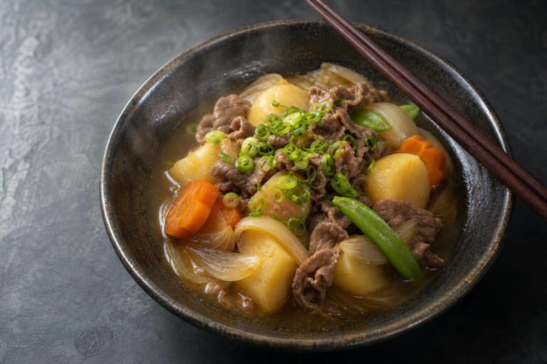 Nikujaga, a Japanese beef and potato stew, in a dark ceramic bowl with tender beef, soft potatoes, carrot, and snow peas in a dashi broth topped with scallions