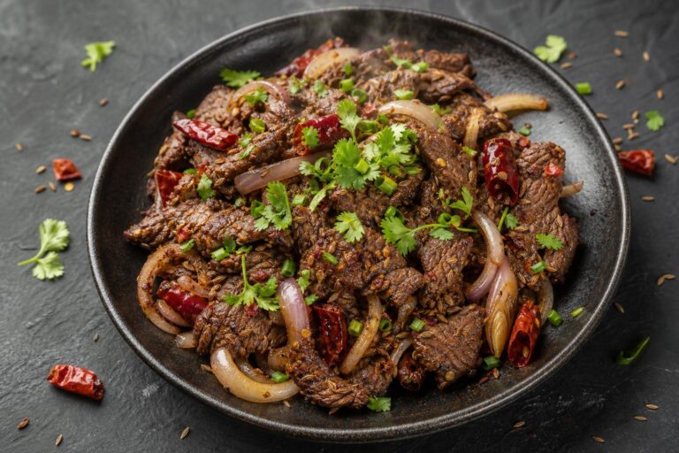 Overhead of Xinjiang cumin beef, charred sliced flank steak coated in whole cumin and chili with onion and dried red chilies, cilantro and scallion on top, in a dark bowl on a slate surface