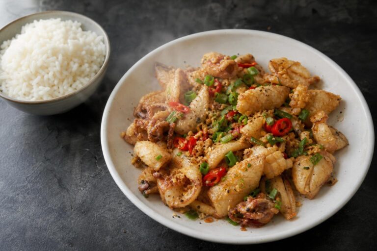 Chinese salt and pepper squid with sliced red chili and scallion in a white bowl beside jasmine rice on dark slate
