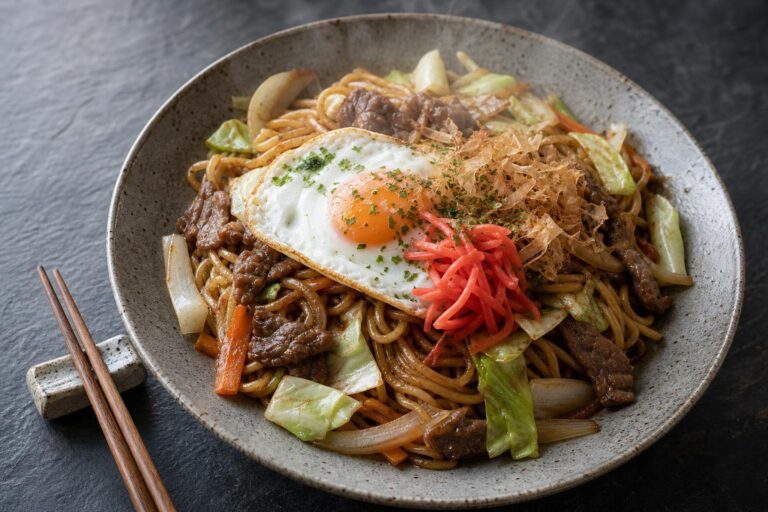 Beef yakisoba with fried egg, pickled ginger and bonito flakes in a ceramic bowl on dark slate
