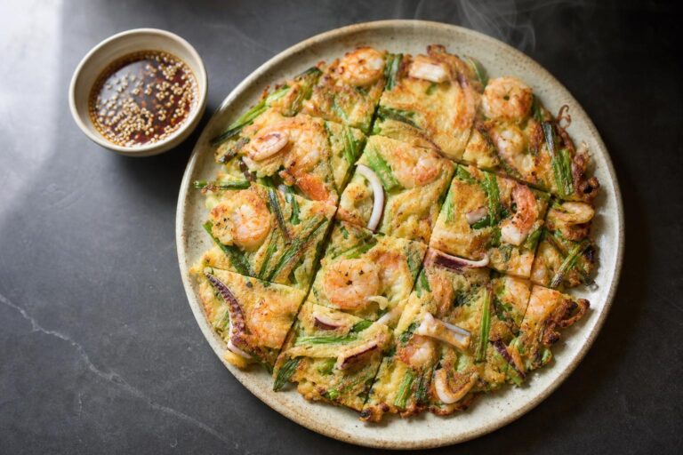 Crispy Korean seafood pancake cut into squares with shrimp, squid and scallions beside a bowl of soy dipping sauce on dark slate