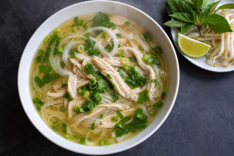 Bowl of Vietnamese chicken pho with shredded chicken, rice noodles, scallions and cilantro beside bean sprouts, basil and lime on dark slate